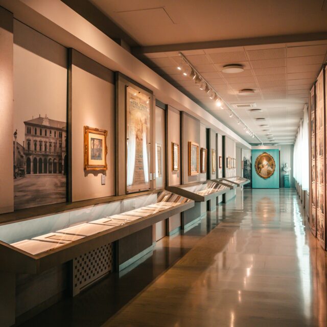 fotografia degli spazi della galleria beltrame nel palazzo del comune di Arzignano. sulla sinistra c'è la scritta "Galleria Beltrame", setuita da quadri in cornice appesi alla parete e una lunga teca orizzontale che contiene i fogli originali del giornale "La Domenica del Corriere" con i disegni di Achille Beltrame