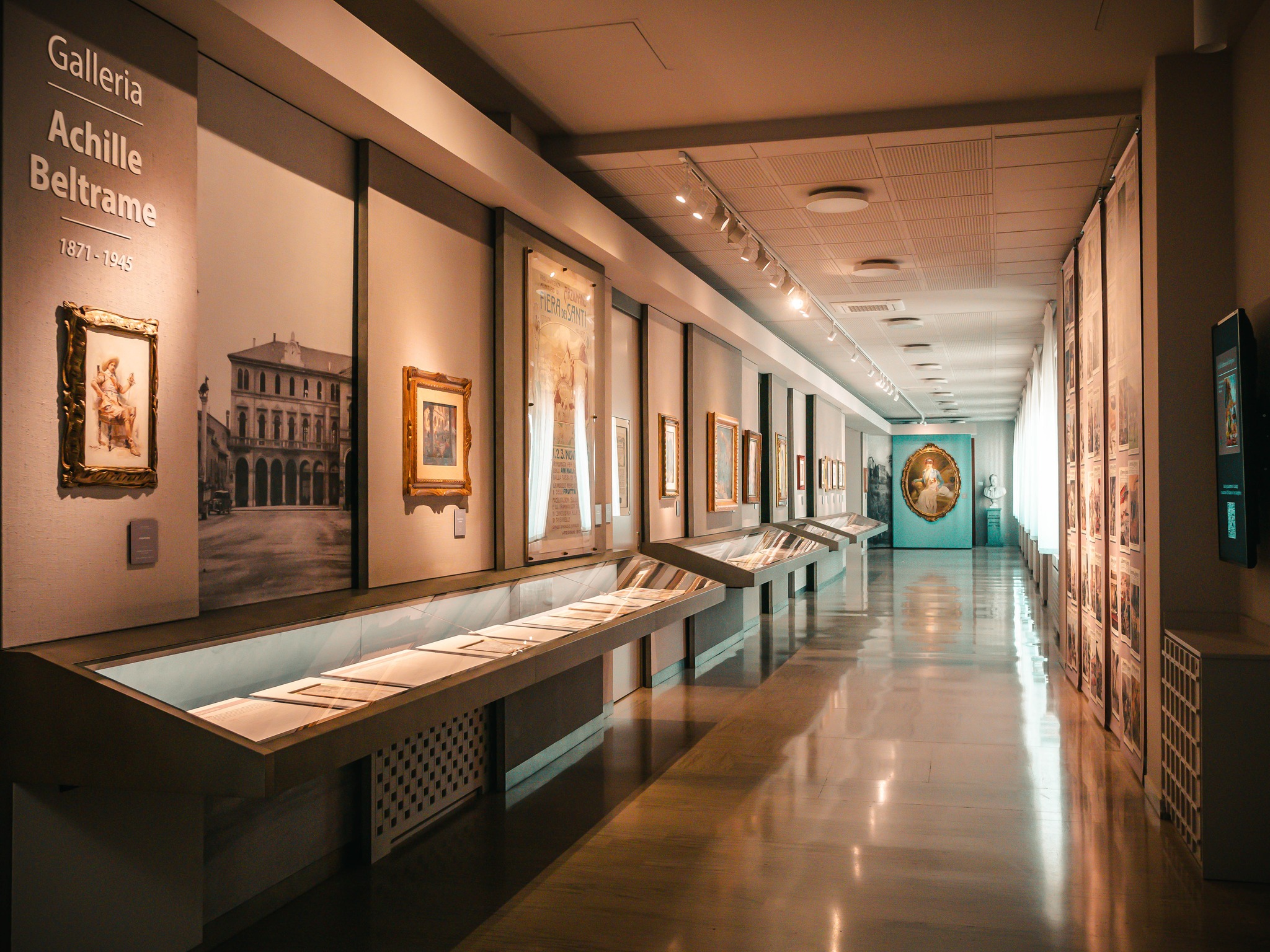 fotografia degli spazi della galleria beltrame nel palazzo del comune di Arzignano. sulla sinistra c'è la scritta "Galleria Beltrame", setuita da quadri in cornice appesi alla parete e una lunga teca orizzontale che contiene i fogli originali del giornale "La Domenica del Corriere" con i disegni di Achille Beltrame